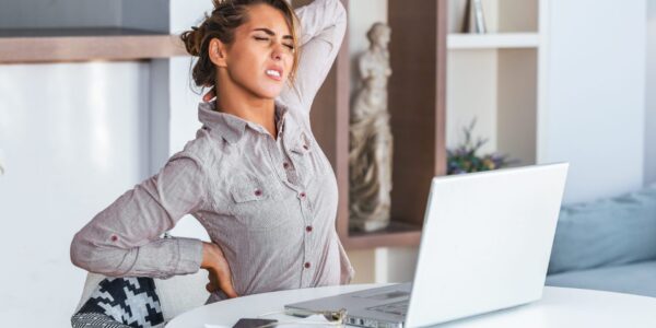 Portrait of young stressed woman sitting at home office desk in front of laptop, touching aching neck with pained expression, suffering from neck pain after working on pc